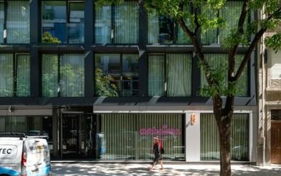 Modern hotel building with dark metal frame and large glass windows. Trees in front cast shadows on the facade. A person walks past the entrance.