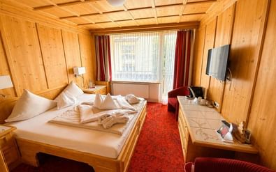 Cozy double room with pine wood paneling, white bedding, red carpet, TV, and window with red curtains at Hotel Latini in Zell am See.