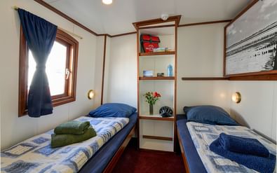 Twin cabin on ship Elizabeth with two single beds, blue bedding, towels, wooden shelving unit, porthole window with curtain, and maritime photograph.