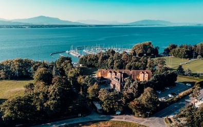 Luftaufnahme des Hotels La Barcarolle in Prangins, ein rotes Backsteingebäude umgeben von Bäumen mit Yachthafen und Genfersee im Hintergrund.