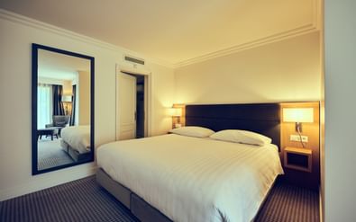 Modernes Hotel-Doppelzimmer mit weißer Bettwäsche, dunklem Kopfteil, Nachttischlampen, großem Spiegel und gemustertem Teppich im Hotel La Barcarolle in Prangins.