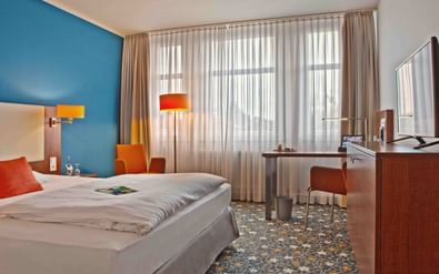 Modernes Hotelzimmer mit blauer Akzentwand, Doppelbett, orangen Stühlen, Schreibtisch, TV und großen Fenstern mit Gardinen im Best Western Hotel President Bonn.