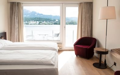 Helles Hotelzimmer mit Doppelbett, bordeauxrotem Sessel und Balkontür mit Blick auf See und Berge in Luzern.