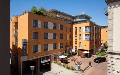 Orange-colored Best Western Hotel building in Bamberg with courtyard terrace, outdoor seating, and modern architecture under blue sky.