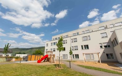 Modern white JUFA Hotel building in Bernkastel-Kues with playground featuring red slide and climbing frame on lawn, hills in background.