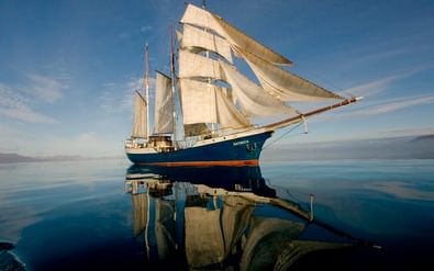 Blau-rumpfiges Segelschiff Antigua mit weißen Segeln, spiegelt sich in ruhigem Wasser unter klarem Himmel mit Bergen am Horizont.