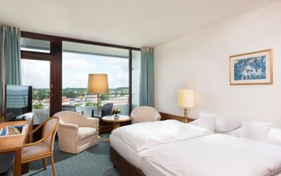 Helles Hotelzimmer mit zwei Einzelbetten, Sitzbereich, Schreibtisch und Balkon mit Blick auf Travemünde. Weiße Bettwäsche und moderne Möbel.