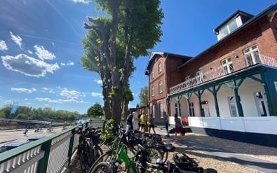 Cyclists standing near parked bicycles in front of a red brick hotel with green veranda. Blue sky and tall trees visible in the background.