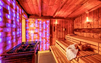 Finnish sauna with wooden benches and colorful LED salt wall illuminated in purple and orange. A sauna stove is visible in the foreground.