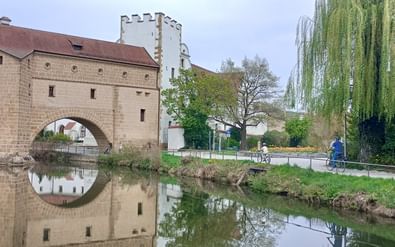 Steinbrücke mit zwei Bögen spiegelt sich im Fluss Vils in Amberg. Radfahrer auf Weg am Wasser, Weide rechts, mittelalterlicher Turm sichtbar.