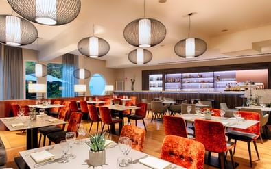 Modern restaurant interior at Welcome Hotel Marburg with orange velvet chairs, white tables, woven pendant lights, and open kitchen.