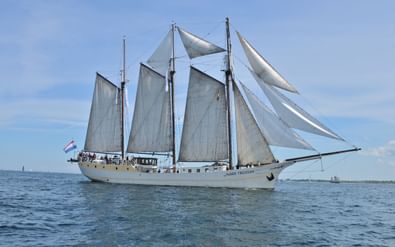 Dreimaster-Segelschiff Mare Frisium mit weißen Segeln auf ruhigem blauen Wasser unter klarem Himmel mit niederländischer Flagge.