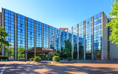Modern glass facade of Sheraton Essen Hotel with blue reflective windows, covered entrance, and landscaped plaza with trees and shrubs.