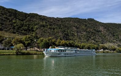 MS Switzerland Flusskreuzfahrtschiff mit weißem Rumpf und blauem Oberdeck auf ruhigem grünem Wasser vor bewaldeten Hügeln und Dorf.