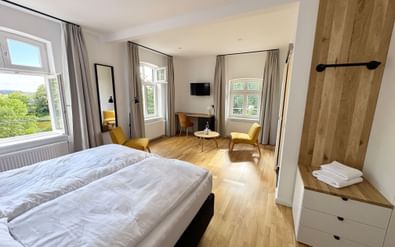 Modern hotel room with double bed, wooden flooring, yellow armchairs, large windows with gray curtains, wall-mounted TV, and wooden wardrobe.