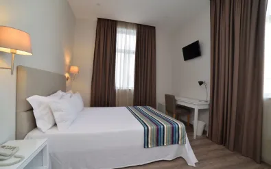 Modern hotel room at Porto Domus Hotel with white double bed, striped runner, brown curtains, wall-mounted TV, desk, and wooden flooring.