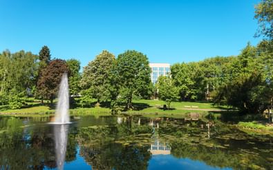 Park pond with fountain reflecting trees and the Sheraton Essen hotel building in the background under a clear blue sky.