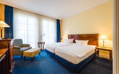 Helles Hotelzimmer mit zwei Einzelbetten, Holzmöbeln, blauem Teppich, gestreiftem Sessel und großen Fenstern mit blau-goldenen Vorhängen in der Villa Geyerswörth.