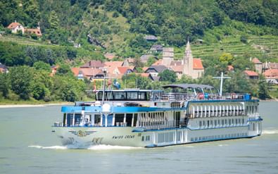 MS Swiss Crown river cruise ship sailing on calm water with a picturesque village featuring red-roofed houses and a church tower on hillside.