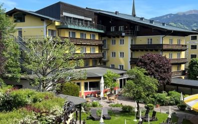 Yellow and brown alpine hotel with balconies and garden terrace with sun loungers. Church spire and mountains visible in background under blue sky.