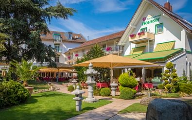 Exterior view of Gartenhotel Heusser with landscaped garden, Japanese stone lanterns, outdoor terrace with yellow awnings, and white building facade.
