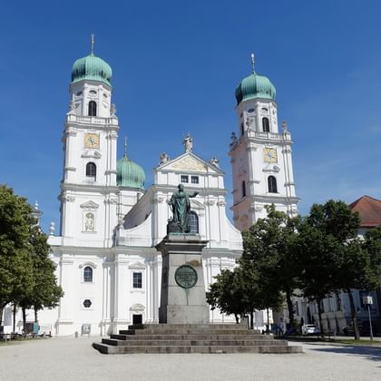 White baroque cathedral in Passau with twin green copper domes and clock towers. A bronze statue stands on a pedestal in the square.