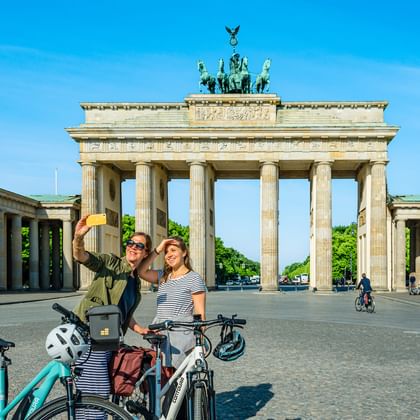 Zwei Radfahrerinnen machen ein Selfie vor dem Brandenburger Tor in Berlin. Sie stehen mit ihren Fahrrädern auf dem Kopfsteinpflaster.