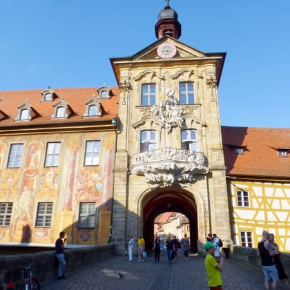 Ornate baroque city gate in Bamberg with decorated tower, painted facades, and red tile roofs. Tourists walk through the arched entrance.
