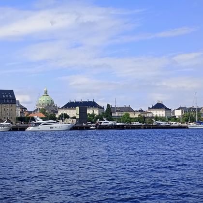 Blick über blaues Hafenwasser in Kopenhagen mit Booten, historischen Gebäuden und einer Kirche mit grüner Kuppel unter teils bewölktem Himmel.
