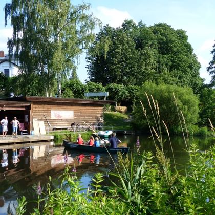 Canoe with passengers departing from wooden boathouse on calm waterway in Billetal. Green vegetation and trees line the water's edge.