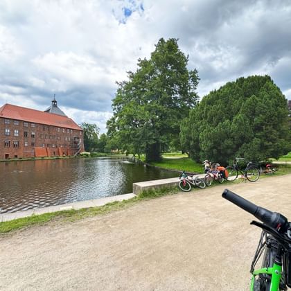 Red brick Winsen Castle beside a moat with cyclists resting on a gravel path. Green trees and cloudy sky frame the historic building.