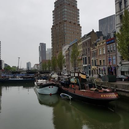 Historic boats moored in Rotterdam harbor with tall brown tower and traditional Dutch buildings along the waterfront under overcast sky.