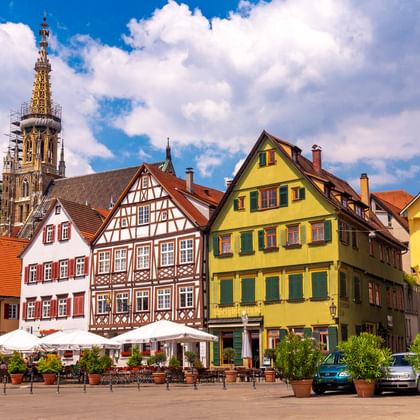 Esslinger Marktplatz mit bunten Fachwerkhäusern und gelben Gebäuden. Kirchturm ragt im Hintergrund unter blauem Himmel mit Wolken empor.