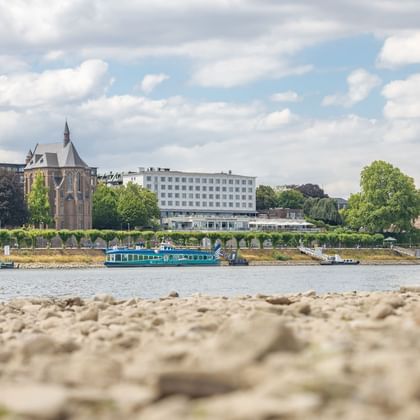 View across the Rhine River in Bonn showing a historic church with spire, modern white buildings, and a blue boat moored at the riverbank.