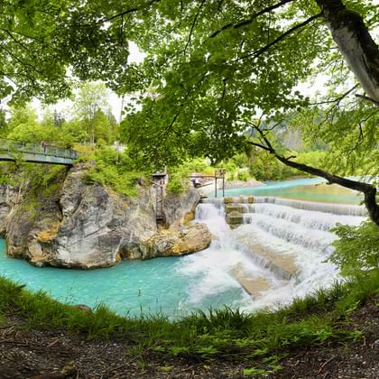 Scenic turquoise river with stepped waterfall flowing through rocky landscape. Large trees frame the scene with a wooden bridge visible.
