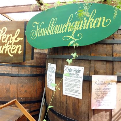 Traditionelle Holzfässer für Spreewälder Gurken mit grünem Schild 'Knoblauchgurken' und braunem Schild 'Senf Gurken' sowie Infoblättern.
