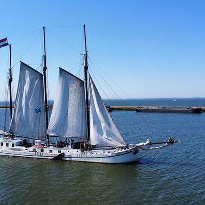 Zweimaster-Segelschiff Elizabeth mit weißen Segeln auf ruhigem blauen Wasser unter klarem Himmel, mit Küstenlandschaft im Hintergrund.