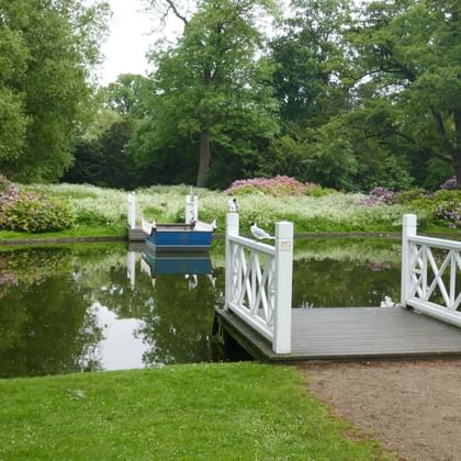 Weißer Holzsteg mit dekorativen Geländern in einem Teich im Frederiksberg Park, Kopenhagen. Ein blaues Ruderboot liegt in der Nähe, umgeben von üppigem Grün und blühenden Sträuchern.