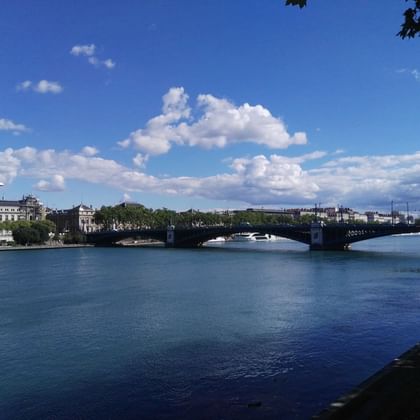 Wide view of a stone bridge crossing the Rhône River in Lyon under blue sky with white clouds. Historic buildings line the left riverbank.