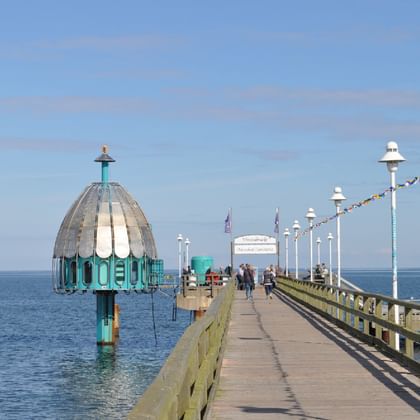 Hölzerne Seebrücke in die Ostsee mit charakteristischer türkisfarbener Tauchgondel und weißen Laternen unter blauem Himmel.