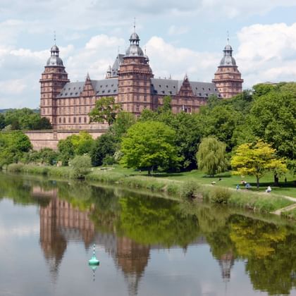 Johannisburg Castle in Aschaffenburg with red sandstone towers reflected in the calm waters of River Main, surrounded by lush green trees.