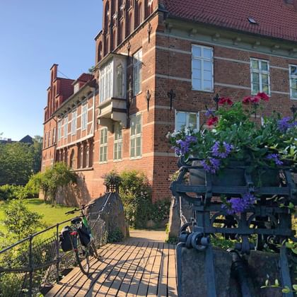 Red brick Bergedorf Castle with wooden bridge entrance decorated with purple and pink flowers. A bicycle leans against the bridge railing.