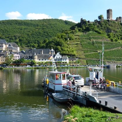 Car ferry on the Moselle River in Beilstein with historic village, vineyards on hillsides, and medieval castle ruins on the hilltop.