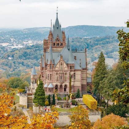 Schloss Drachenburg mit verzierten Türmen und Spitzen, umgeben von Herbstlaub. Das Rheintal und Hügel sind im Hintergrund sichtbar.