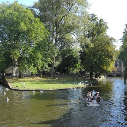 Park mit Kanal und Boot in Brügge Malerischer Park in Brügge mit einem Kanal. Ein kleines Boot mit Passagieren fährt durch das Wasser, während Schwäne in der Nähe schwimmen. Üppige grüne Bäume umgeben das Gebiet.
