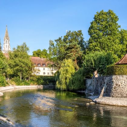 Konstanz city garden with river, stone bridge, and Konstanz Cathedral's twin spires visible in the background among green trees under blue sky.