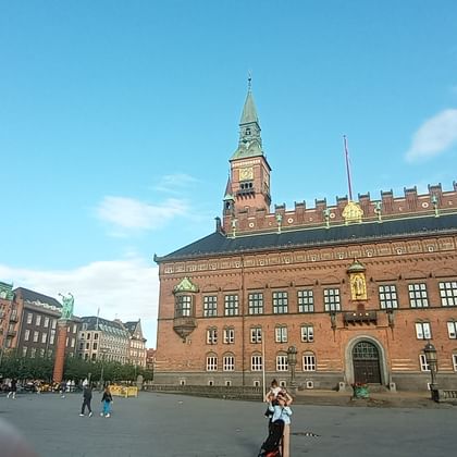 Copenhagen City Hall, a red brick building with clock tower, at Rådhuspladsen square under blue sky with pedestrians in the foreground.