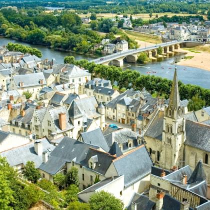 Aerial view of Chinon, France showing medieval stone buildings with slate roofs, church spire, and stone bridge crossing the Loire River.