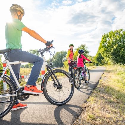 Family of cyclists on a paved bike path in Thuringia. Adult in green shirt and helmet rides in foreground, with two children in colorful clothing behind.