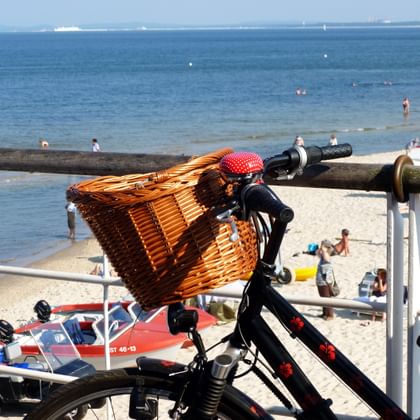 Black bicycle with wicker basket parked at Usedom beach railing, overlooking sandy beach with beachgoers and blue Baltic Sea.